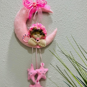 May include: A pink felt baby mobile featuring a crescent moon with a sleeping baby doll wearing a floral bonnet. The mobile includes three pink star-shaped ornaments with closed eyes, and a pink bow.