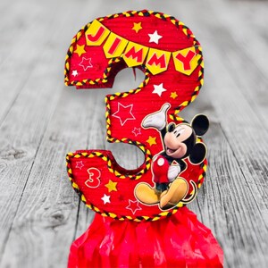 May include: Red and yellow Mickey Mouse themed pi&ntilde;ata shaped like the number 3. The pi&ntilde;ata has a banner that says "JIMMY" and features Mickey Mouse in the centre.