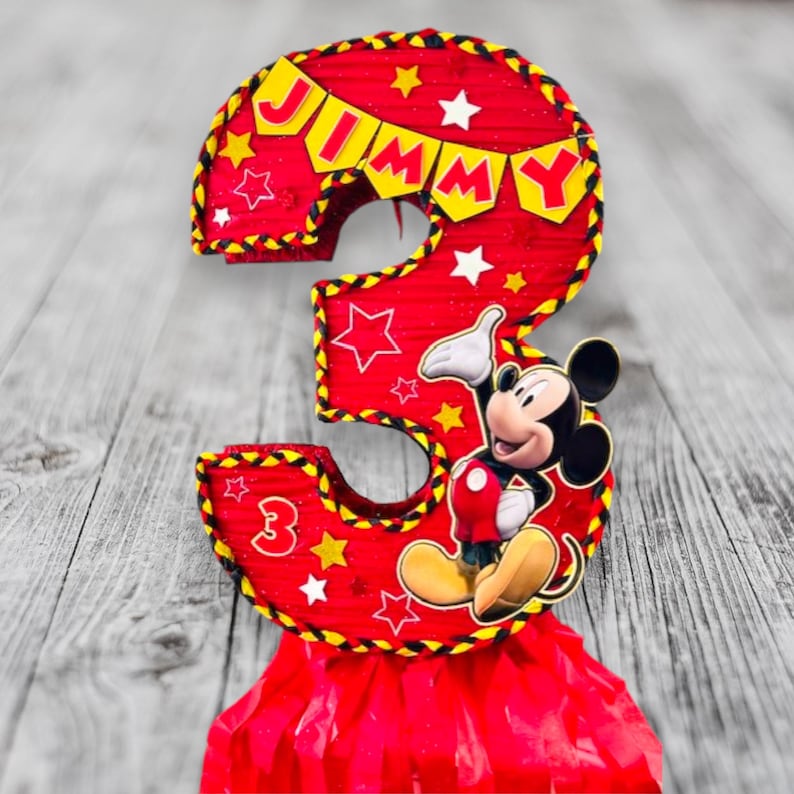 May include: Red and yellow Mickey Mouse themed pi&ntilde;ata shaped like the number 3. The pi&ntilde;ata has a banner that says "JIMMY" and features Mickey Mouse in the centre.