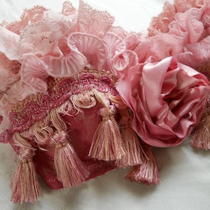 May include: Close-up of a decorative textile with layers of pink lace, ruffles, and tassels. A large, pink satin rose is featured on the right side. The textile has a vintage, romantic aesthetic.