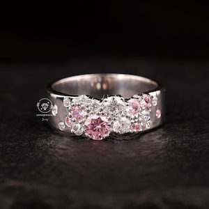 May include: A silver ring with a floral design. The ring features a large pink gemstone in the centre, surrounded by smaller pink and clear gemstones. The band has a textured surface with small, round accents. The ring is set against a dark background.