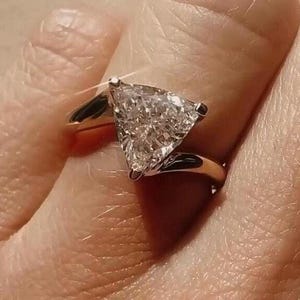May include: A gold ring with a triangular diamond set in a prong setting. The ring is on a finger.