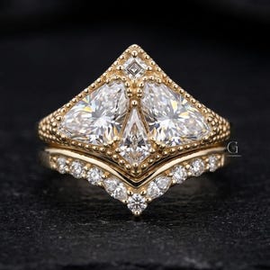 May include: A two-piece gold ring set. The top ring features a triangular design with three large, clear stones. The bottom ring has a curved band with smaller, clear stones. The rings are set against a dark background.