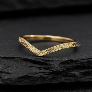 May include: A gold-colored chevron ring with an engraved floral pattern. The ring is set against a dark background, highlighting its delicate design. The ring's V-shape and intricate details make it a unique piece of jewelry.