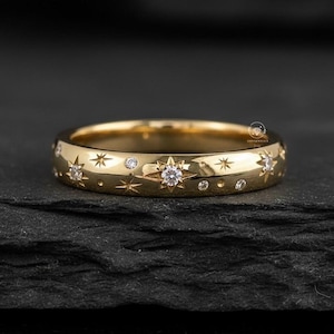 May include: A gold ring with a celestial design. The ring features star and dot engravings, with small, clear gemstones set into the band. The ring is displayed on a dark, textured surface.