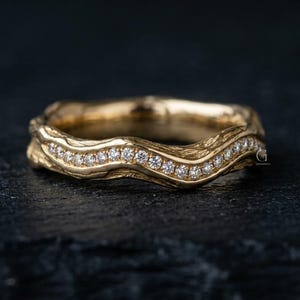 Può includere: Un anello in oro con una texture organica ondulata, caratterizzato da una fila di piccole pietre rotonde e trasparenti incastonate al centro. La fascia ha una finitura lucida e una forma sinuosa che segue il profilo delle pietre.