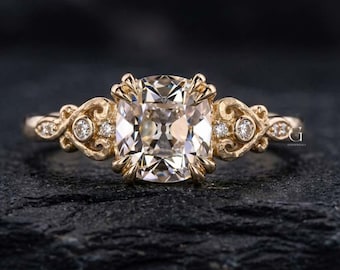 May include: A gold ring featuring a large cushion-cut center stone held by four prongs. The band displays intricate scrollwork detailing on both sides, accented with small round diamonds embedded within the metalwork, creating a detailed design.