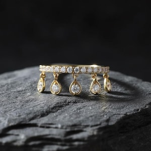 May include: A gold ring with a band encrusted with small, round diamonds. The ring features teardrop-shaped diamond pendants hanging from the band. The ring is displayed on a dark gray, textured surface.