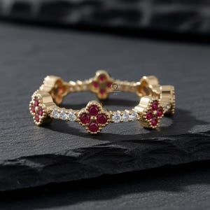 May include: A gold ring with a repeating clover design. Each clover is made of small, round, red gemstones. The ring also features small, round, white gemstones between the clovers. The ring is set against a dark background.