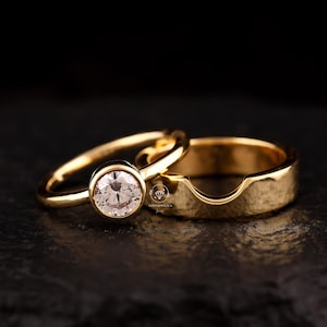 May include: Two gold wedding rings. One ring features a round, clear gemstone set in a bezel. The other ring is a simple gold band with a curved indentation. The rings are displayed on a dark surface.