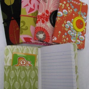 以下が含まれることがあります： さまざまな花柄や葉柄の生地で覆われたノートのコレクション。1冊のノートが開いており、罫線入りの紙とギフトカードが入ったポケットが見えます。ノートは緑、ピンク、オレンジ、黒の色合いで、ゴムやボタンで留められています。