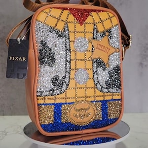 Può includere: Una borsa a tracolla in pelle marrone con un design di Woody di Toy Story realizzato con strass. La borsa ha una tracolla marrone e un'etichetta che dice "PIXAR". La borsa ha anche un distintivo a stella da sceriffo che dice "SHERIFF".