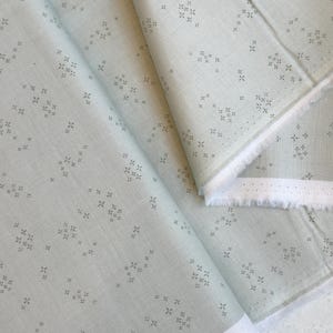 May include: A light green fabric with a repeating pattern of small, gray cross-like shapes. The fabric is folded, revealing a white edge with small perforations. The material appears to be cotton or linen.