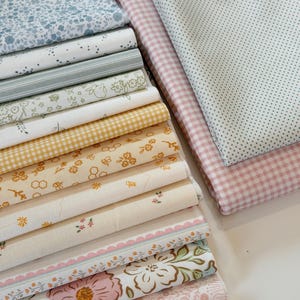 May include: A stack of folded fabric squares featuring various patterns including blue floral, green stripes, yellow gingham, honeycombs, and large pink flowers. To the side, two additional folded fabrics show pink gingham and a white dotted pattern.