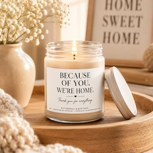 Könnte beinhalten: Eine Kerze im klaren Glas mit brennendem Docht und einem weißen Etikett mit dem schwarzen Text "BECAUSE OF YOU, WE'RE HOME. Thank you for everything". Die Kerze enthält hellbeiges Wachs und hat einen separaten weißen Metalldeckel auf einem Holztablett.