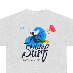 May include: Light gray t-shirt featuring a colorful graphic of a person kitesurfing. The design includes a blue wave, a pink kite, and the words "Kite Surf" with "La Ventana, MX" below. Two birds are also included.