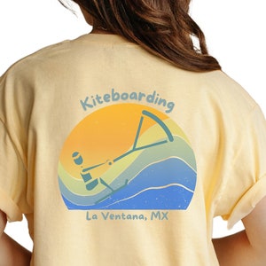 May include: A yellow t-shirt with a retro-style graphic on the back. The graphic features a person kiteboarding with a sunset and waves in the background. The text "Kiteboarding La Ventana, MX" is below the graphic.