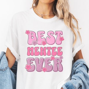 May include: White t-shirt with the text "BEST MENTEE EVER" in pink and purple bubble letters, decorated with small flower graphics. The shirt is worn with a denim jacket.