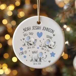 Op de afbeelding: Een witte keramische kerstbal met een gouden draad, met de naam "NOAH JOSIAH JOHNSON" en baby-mijlpalen: "ROLL OVER", "CRAWL" en "WALK". Bevat data en olifantenillustraties. De bal hangt aan een kerstboom.