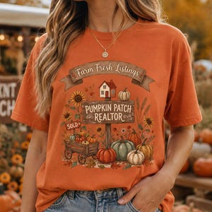 May include: An orange short-sleeve t-shirt featuring a graphic print with the text Farm Fresh Listings and PUMPKIN PATCH REALTOR. The design includes a small house, a wooden wagon, various pumpkins, sunflowers, and a sign labelled SOLD.