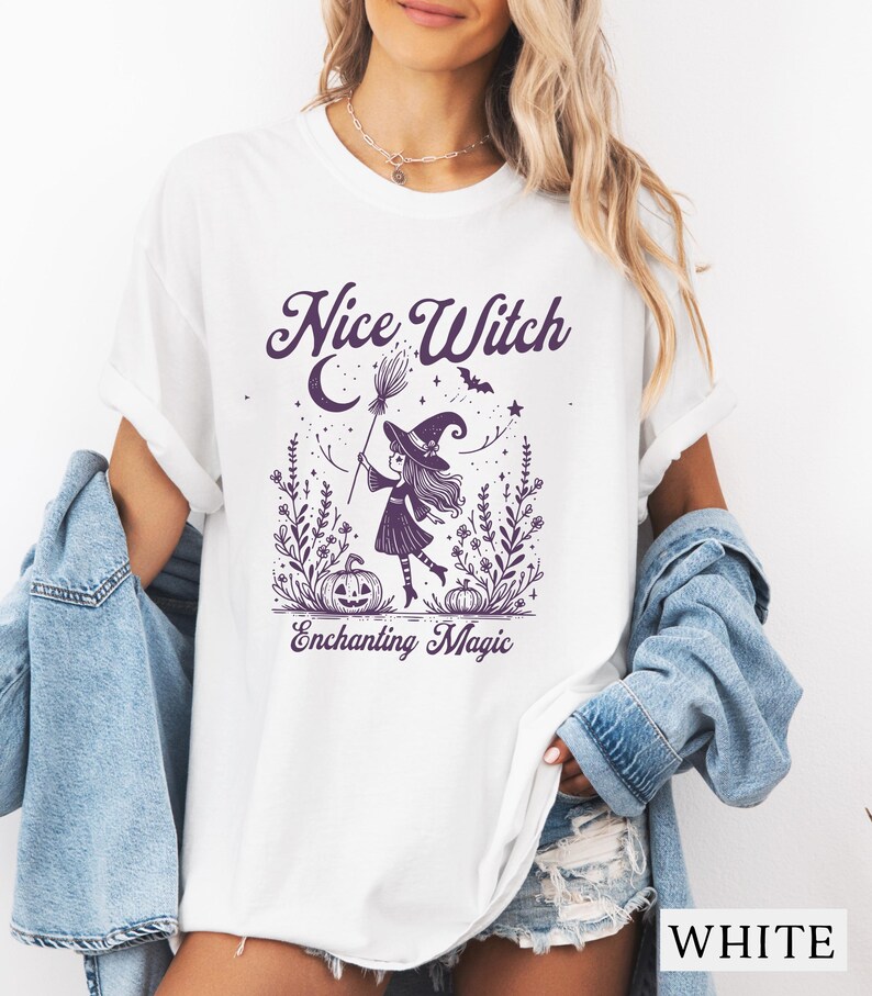 May include: White t-shirt with a purple graphic of a witch holding a broom, surrounded by a pumpkin, flowers, and the text "Nice Witch Enchanting Magic". The shirt has short sleeves.