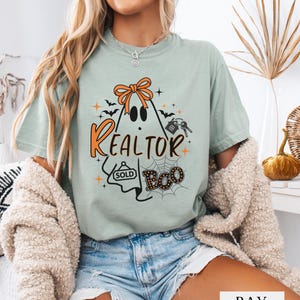 Könnte beinhalten: Ein salbeigrünes Kurzarm-T-Shirt mit einer Grafik eines Geistes, der eine orangefarbene Schleife trägt. Das Design enthält den Text REALTOR und BOO sowie Illustrationen von Fledermäusen, einem Spinnennetz, einem Hausschlüssel und einem SOLD-Schild.