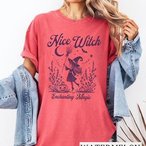 May include: Watermelon-colored t-shirt with the text "Nice Witch" above an illustration of a witch with a broom, a crescent moon, and pumpkins. The text "Enchanting Magic" is below the illustration. The shirt is a comfortable, casual style.