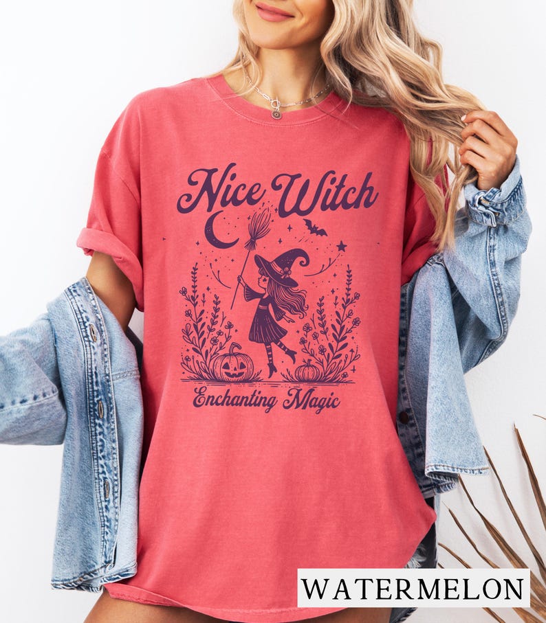 May include: Watermelon-colored t-shirt with the text "Nice Witch" above an illustration of a witch with a broom, a crescent moon, and pumpkins. The text "Enchanting Magic" is below the illustration. The shirt is a comfortable, casual style.