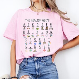 May include: A pink t-shirt with a white graphic print of the alphabet with real estate terms and icons. The text reads "THE REALTOR ABC'S".