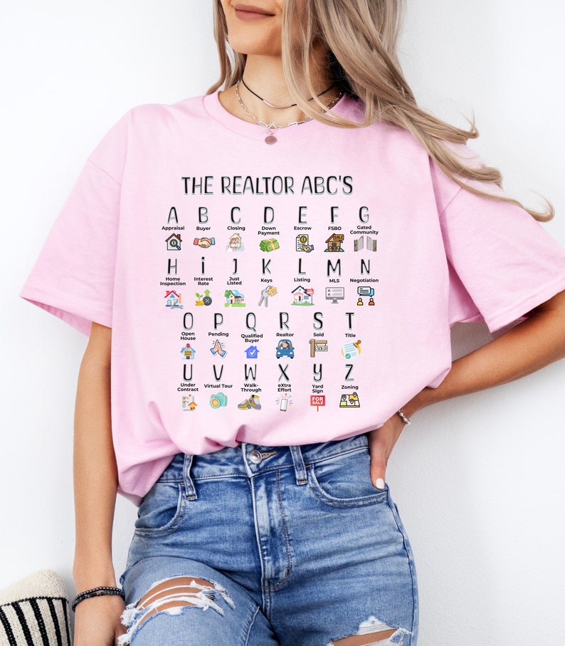 May include: A pink t-shirt with a white graphic print of the alphabet with real estate terms and icons. The text reads "THE REALTOR ABC'S".