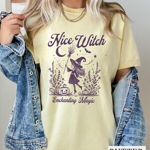 May include: Butter-colored t-shirt with a purple graphic of a witch holding a broom, a crescent moon, and the words "Nice Witch" and "Enchanting Magic". The witch is wearing a hat and standing in a field of flowers and pumpkins. The shirt is worn with a denim jacket.