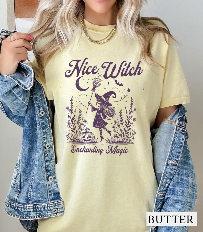 May include: Butter-colored t-shirt with a purple graphic of a witch holding a broom, a crescent moon, and the words "Nice Witch" and "Enchanting Magic". The witch is wearing a hat and standing in a field of flowers and pumpkins. The shirt is worn with a denim jacket.