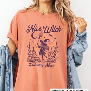 May include: Terracotta-colored t-shirt with a purple graphic of a witch holding a broom, with the words "Nice Witch" and "Enchanting Magic". The witch is wearing a hat and standing in a field with pumpkins and stars.
