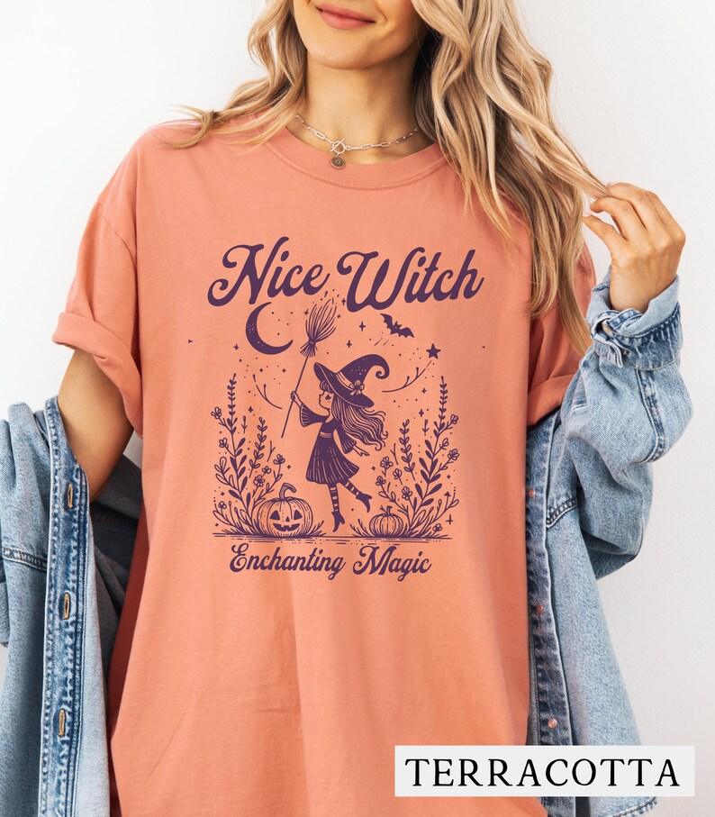 May include: Terracotta-colored t-shirt with a purple graphic of a witch holding a broom, with the words "Nice Witch" and "Enchanting Magic". The witch is wearing a hat and standing in a field with pumpkins and stars.