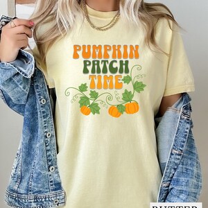 May include: A pale yellow short-sleeve t-shirt featuring the text PUMPKIN PATCH TIME in orange and green letters. The design includes green vines with leaves and three orange pumpkins. A blue denim jacket is worn over the t-shirt.
