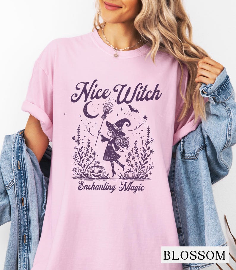 May include: A light pink t-shirt with the words "Nice Witch" and "Enchanting Magic" printed in dark purple. The graphic features a witch with a broom, pumpkins, and floral elements. The word "BLOSSOM" is printed at the bottom.