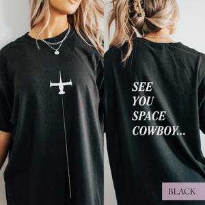 May include: Black t-shirt featuring a white graphic of a spaceship and a vertical line. The back of the shirt displays the text "SEE YOU SPACE COWBOY..." in white. The bottom right corner of the back of the shirt has the word "BLACK".
