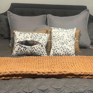 May include: A bed is made with gray quilted bedding, gray pillows, and decorative pillows. Two pillows have a white leaf pattern. A chunky, orange blanket is at the foot of the bed. A pillow has a pocket.