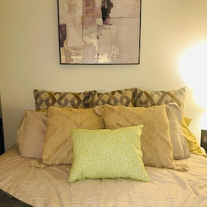 May include: A bed with a beige comforter and several pillows. The pillows include patterned, textured, and solid colors. A green floral pillow is in the center. A clock, artwork, and a lamp are also visible.