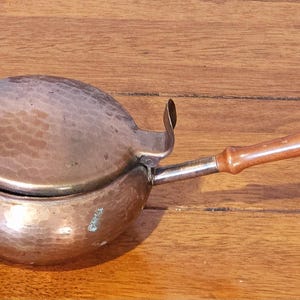 Op de afbeelding: Een koperkleurige, gehamerde metalen kookpot met een deksel en een houten handvat. De pot heeft een afgeronde vorm en een kleine tuit. De handgreep is bevestigd met zilverkleurig metaal. De pot staat op een houten oppervlak.