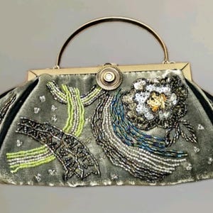May include: A vintage-style handbag in a muted green colour, adorned with intricate beadwork. The bag features a silver handle and clasp, with floral and abstract designs in silver, white, and green beads. The bag's edges are trimmed with black fabric.