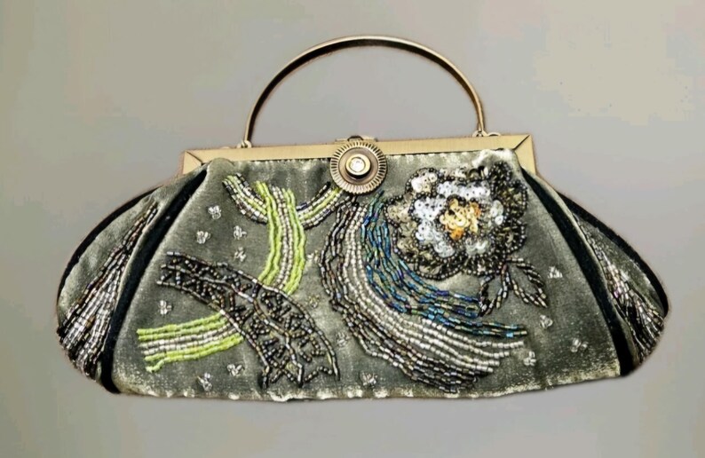 May include: A vintage-style handbag in a muted green colour, adorned with intricate beadwork. The bag features a silver handle and clasp, with floral and abstract designs in silver, white, and green beads. The bag's edges are trimmed with black fabric.