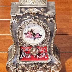 Könnte beinhalten: Eine antike silberfarbene Kaminuhr mit marmorähnlichem Finish. Das Zifferblatt zeigt cherubische Figuren und römische Ziffern. Die Uhr hat eine verzierte Detaillierung, einen roten Akzent und dekorative Füße.