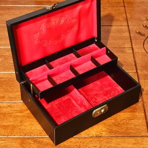 Puede incluir: Caja de joyería negra abierta con forro de terciopelo rojo y múltiples compartimentos. La caja tiene un cierre dorado y una cerradura. El interior presenta un diseño decorativo en la tapa.
