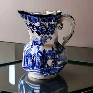 Puede incluir: Una jarra de cerámica azul y blanca con un diseño floral y un asa. La jarra tiene una forma geométrica y está decorada con un patrón floral azul y blanco.