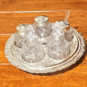 Könnte beinhalten: Ein silberfarbenes Serviertablett mit aufwendigen Details hält fünf klare Glasbehälter mit silbernen Deckeln. Die Gläser sind mit einem strukturierten Muster versehen. Ein Glas hat einen kleinen Löffel. Das Set ist wahrscheinlich zum Servieren von Gewürzen.