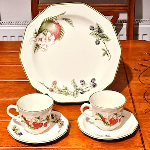 May include: A white ceramic dish set with a large octagonal serving platter, two teacups, and saucers. The set features a green trim and a fruit and floral design with red, green, and white accents.