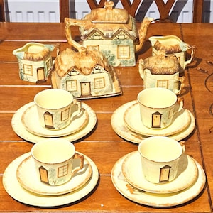 Puede incluir: Un juego de té vintage con diseño de cabaña. El juego incluye una tetera, azucarero, lechera, tazas y platillos. Las piezas son de color crema con detalles en marrón y verde, con un motivo de cabaña.
