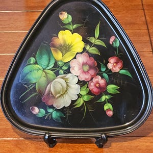 Puede incluir: Bandeja triangular negra con un diseño floral pintado a mano. Las flores son en tonos de amarillo, rosa y blanco, con hojas verdes. La bandeja está soportada por un soporte negro.
