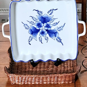 Puede incluir: Una fuente cuadrada de cerámica blanca con un diseño floral azul y bordes festoneados. La fuente tiene dos asas y un borde azul. Descansa sobre una cesta de mimbre marrón.